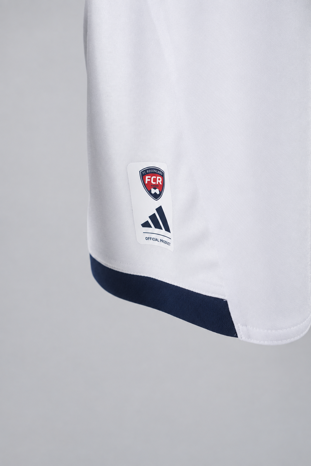 Studio-lit detail shot of the FCR crest and adidas Official Product hem label on the white jersey