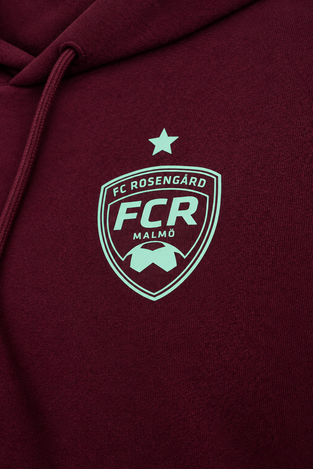 Studio-lit detail shot of the mint FC Rosengård crest on the burgundy hoodie